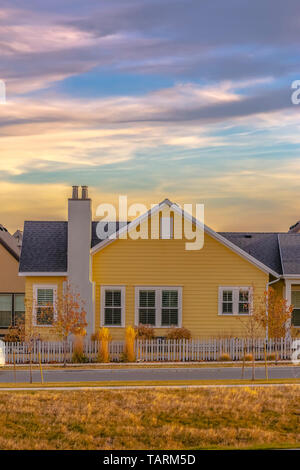 Exterior of a single storey home with yellow wall and dark gray roof. In front of the home is a paved road and grassy terrain under the cloudy blue sk Stock Photo