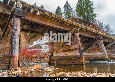 Rustic wooden bridge crossing over a rocky stream with clear shallow water. The bridge overlooks a hill with lush tress against cloudy sky. Stock Photo