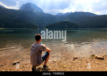 A teenager sits in the early morning on the shore of a mountain lake with a bottle of water and looks at the surface of the water. Stock Photo