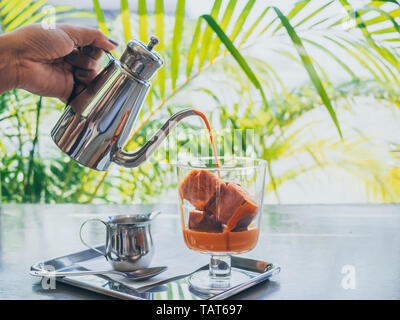 Hand pouring Thai milk tea from stainless steel jug on Thai tea ice cubes in glass with milk in small jug on stainless steel tray on concrete table. Stock Photo