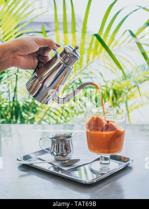 Hand pouring Thai milk tea from stainless steel jug on Thai tea ice cubes in glass with milk in small jug on stainless steel tray on concrete table ve Stock Photo