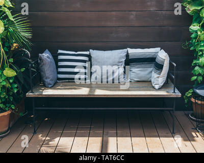 Wooden sofa with pillows between green tropical trees in terra cotta pots on wooden wall background. Outdoor living garden decoration with wooden long Stock Photo