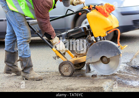 A road maintenance officer in a green reflective vest starts the engine of the gasoline cutter to remove the old asphalt during repairs on the roadway Stock Photo