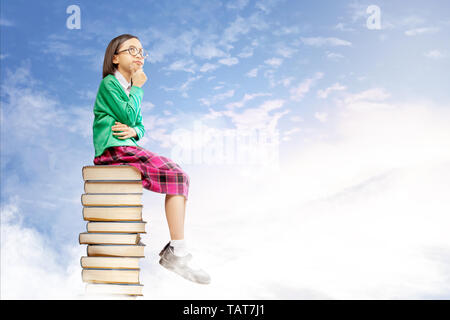 Asian cute girl with glasses think while sitting on the pile of books with blue sky background. Back to School concept Stock Photo