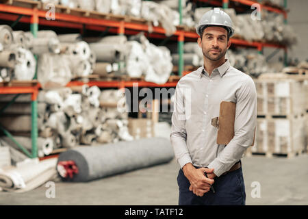 Portrait of a foreman wearing a hardhat and holding a clipboard while standing in a carpet warehouse with stock on shelves in the background Stock Photo
