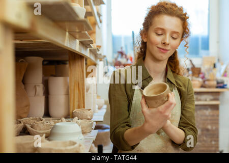 professional workshop. Smiling long-haired curly lady attentively observing clay pot and checking all details Stock Photo