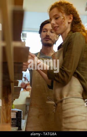 Masterclass in pottery. Smiling ginger woman attentively listening to dark-haired master while checking examples of clay art Stock Photo