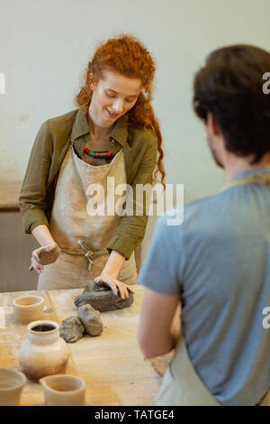 Creating pot. Smiling long-haired student in green shirt pressing piece of clay while learning new techniques in pottery Stock Photo