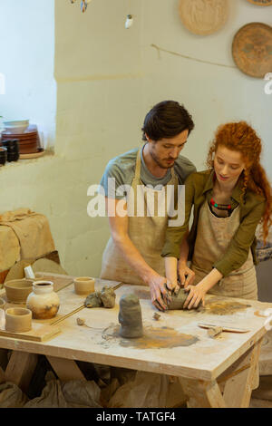 Processing wet clay. Bearded focused teacher explaining his student basics of pottery while staying in equipped workshop Stock Photo