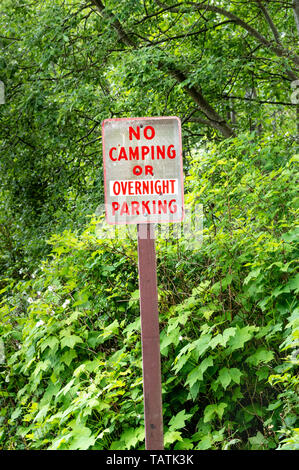 No camping or overnight parking warning sign on post with red lettering on rural road beside green trees. Stock Photo