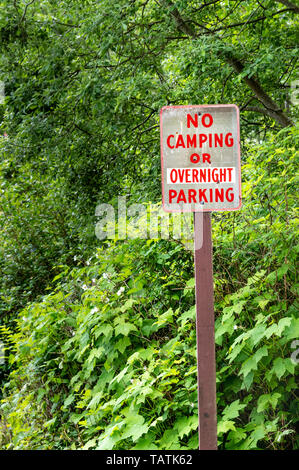 No camping or overnight parking warning sign on post with red lettering on rural road beside green trees. Stock Photo