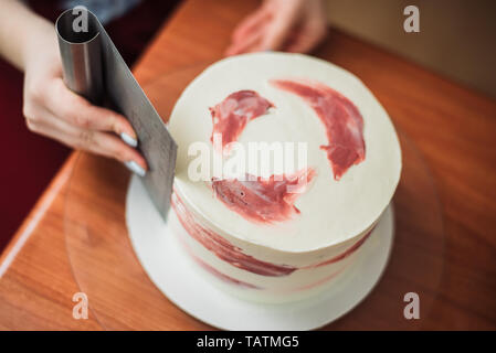 Confectioner using a knife to smooth the sides and top of the cake, close-up top view photo. The concept of home baking, cooking cakes. process of decorating the cake with cream, line the white cream on the cake. Stock Photo