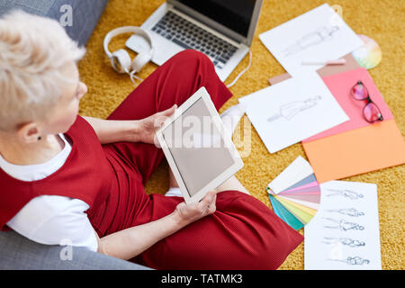 Middle aged female contemporary designer with touchpad sitting on the floor among sketches and other stuff while working over new project Stock Photo