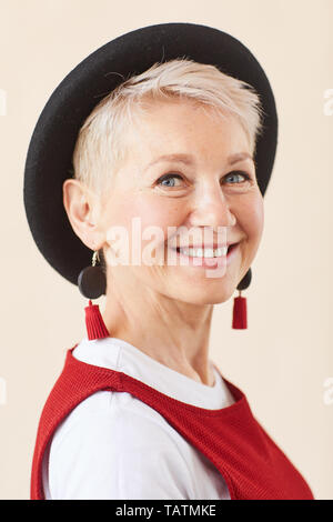 Happy mature blonde female in black hat and smart casual looking at you with toothy smile while posing for camera Stock Photo