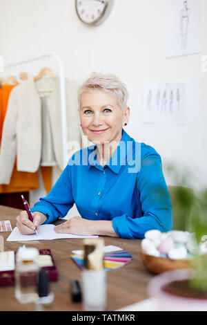 Happy blonde pretty creative fashion designer in blue blouse looking at you while working over new project in studio Stock Photo