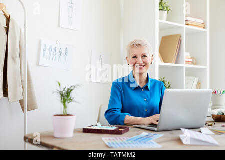 Cheerful mature blonde woman in smart casual sitting by table in front of laptop while surfing through websites for new ideas Stock Photo
