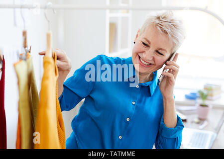 Cheerful blonde woman with smartphone consulting with her friend or daughter while looking for new casualwear in boutique Stock Photo