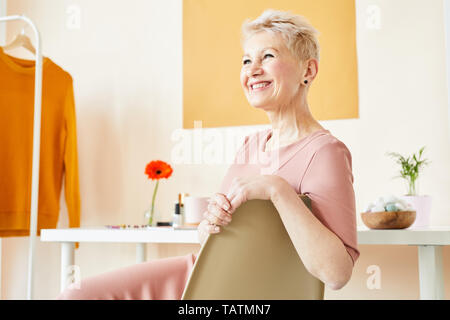 Happy middle-aged blonde female in elegant beige dress sitting on white plastic chair in studio while posing for camera Stock Photo