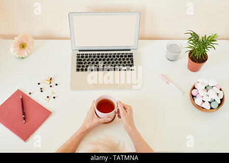 View of human hands holding cup of tea among office supplies, flowers, scented candle and marshmallows on desk Stock Photo