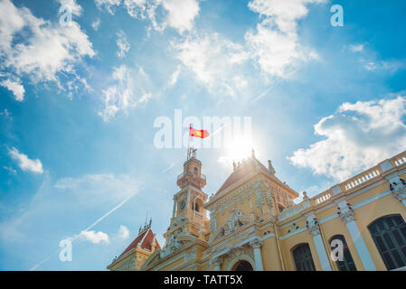 Saigon City Hall with waving Vietnamese flag against blue sky and curly summer clouds, sun, and airplane trail. Stock Photo