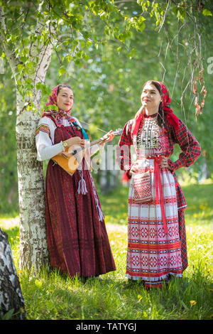 Two smiling woman in traditional russian clothes standing in the forest.  One of them holding balalaika. Vertical shot Stock Photo - Alamy