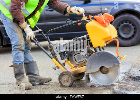 An employee of a road maintenance service in a green reflective vest removes old asphalt with a gasoline cutter during repairs on the roadway. Stock Photo