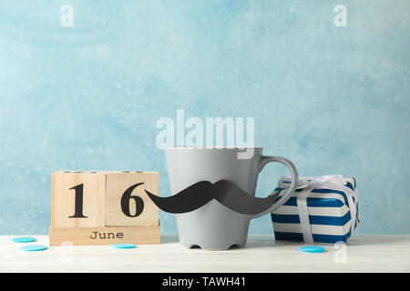 Cup with decorative mustache, gift box and wooden calendar on white table against blue background, space for text Stock Photo
