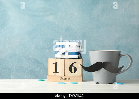 Cup with decorative mustache, gift box and wooden calendar on white table against blue background, space for text Stock Photo