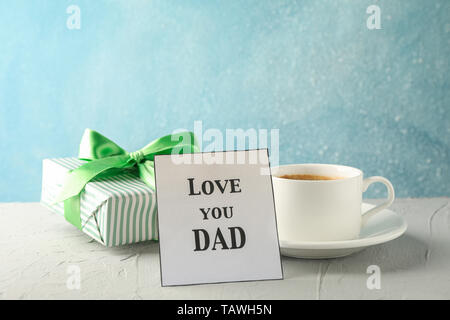 Cup of coffee, gift box with green ribbon and inscription Love you DAD on white table against blue background, space for text Stock Photo