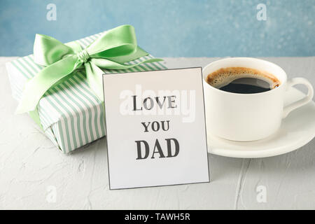 Cup of coffee, gift box with green ribbon and inscription Love you DAD on white table against blue background, space for text Stock Photo