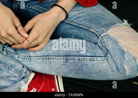 Woman sitting crossed legs with hands in the lap waiting. Ripped denim jeans and red jacket. Touching hands worried/sad Stock Photo