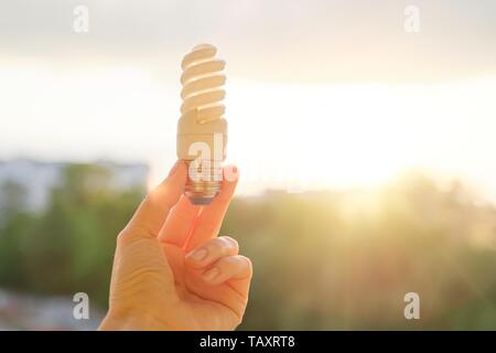 Energy saving fluorescent light bulb, hand holding lamp, evening sunset sky background. Stock Photo