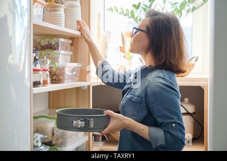 Mature woman in the kitchen pantry with products. Storage wooden stand with kitchenware, products necessary to cook. Stock Photo