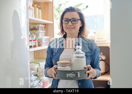 Interior of wooden pantry with products for cooking. Adult woman taking kitchenware and food from the shelves Stock Photo