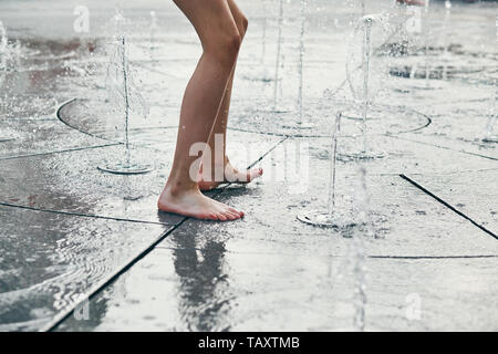 Child standing, jumping, playing in a fountain while hot weather. Closeup of legs, water streams, splashes. Candid people, real moments, authentic sit Stock Photo