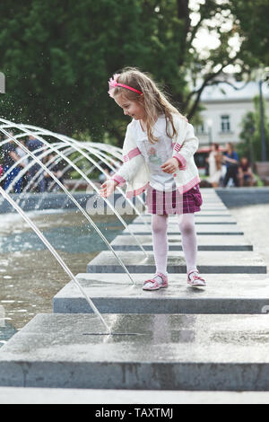 Little happy girl playing with the water in fountain in the center of town. Child catching a water stream going from a fountain. Candid people, real m Stock Photo