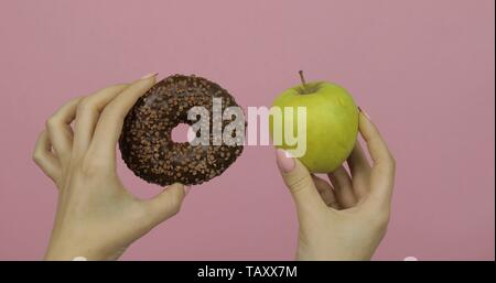 Choice donut against apple. Starting healthy eating or junk food. Girl hands on a pink background holds donut in one hand and apple on other Stock Photo