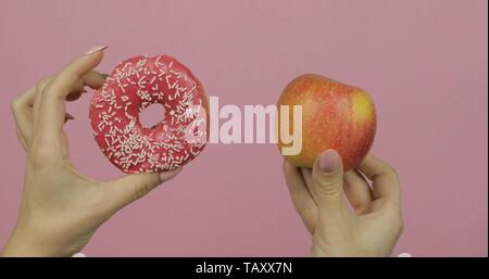 Choice donut against apple. Starting healthy eating or junk food. Girl hands on a pink background holds donut in one hand and apple on other Stock Photo