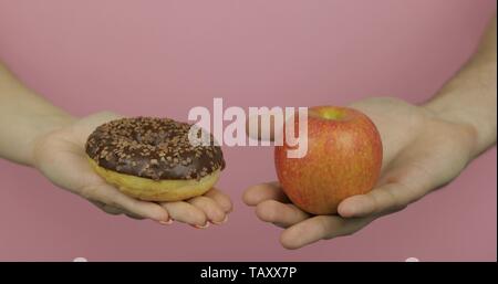 Choice donut against apple. Starting healthy eating or junk food. Hands on a pink background holds donut in one hand and apple on other Stock Photo