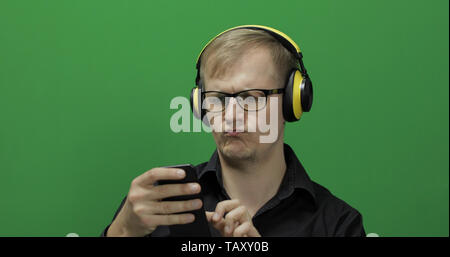 Guy using smart phone in wireless yellow headphones. Portrait of attractive man using mobile phone in headphone while sitting near green screen. Chroma key Stock Photo