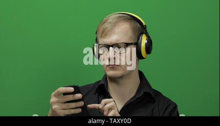 Guy using smart phone in wireless yellow headphones. Portrait of attractive man using mobile phone in headphone while sitting near green screen. Chroma key Stock Photo