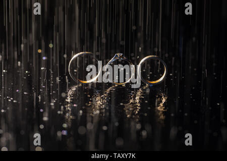 Wedding and engagement rings lying on dark surface shining with light close up macro. Water splashes. Rain Stock Photo