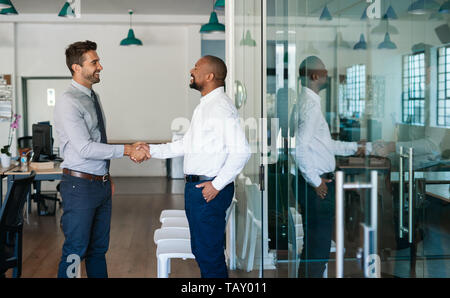 Two diverse businessmen standing in an office laughing and shaking hands together after a successful meeting Stock Photo