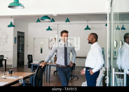 Two diverse young businessmen talking and laughing together while standing in a large modern office Stock Photo