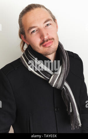 Young Asian man in black coat and woolen scarf stands over white wall. Close up studio portrait Stock Photo