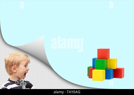 Surprised blond boy standing in an exposed corner is looking at blank page with a pyramid created from colorful wooden cubes. Stock Photo