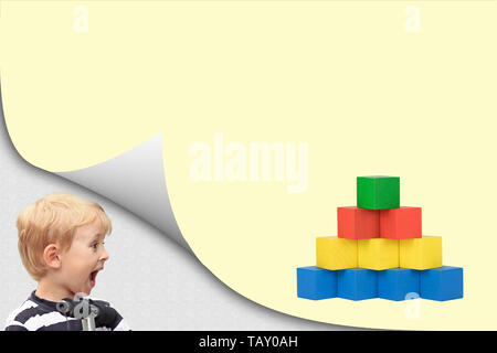 Surprised blond boy in an exposed corner is looking at blank yellow page with a pyramid created from colorful wooden cubes. Stock Photo
