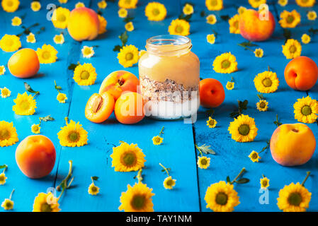 Healthy breakfast with fruit yogurt and oatmeal in a jar, surrounded by yellow flowers and peaches, on a blue table. Diet food context. Stock Photo