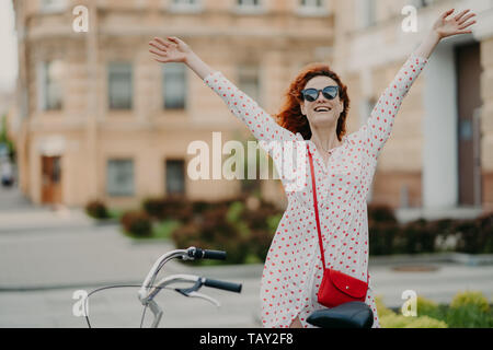 Horizontal outdoor shot of delighted positive red haired woman raises hands, feels free, dressed in summer clothes, rides bicycle on streets, enjoys r Stock Photo