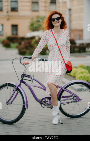 Pleased female has ride bike, dressed in fashionable outfit, smiles happily, stands near bicycle, being photographed in full length against blurred ci Stock Photo